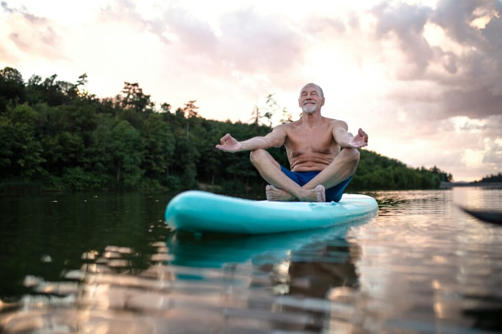Senior man on paddleboard on lake in summer, doing yoga exercise